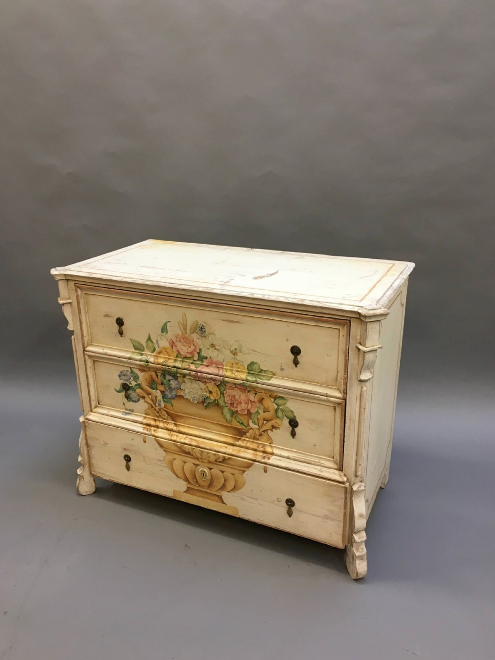 A 19th-century cream painted chest of drawers featuring three long drawers decorated with a central hand-painted motif of a floral urn.