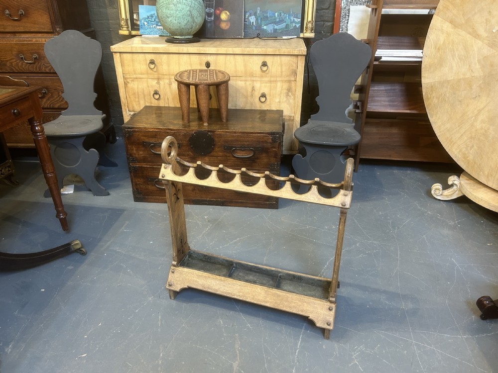 An antique oak umbrella and stick stand featuring a scalloped upper rail, carved ring handles, inlaid side uprights, and a metal drip tray at the base. View X