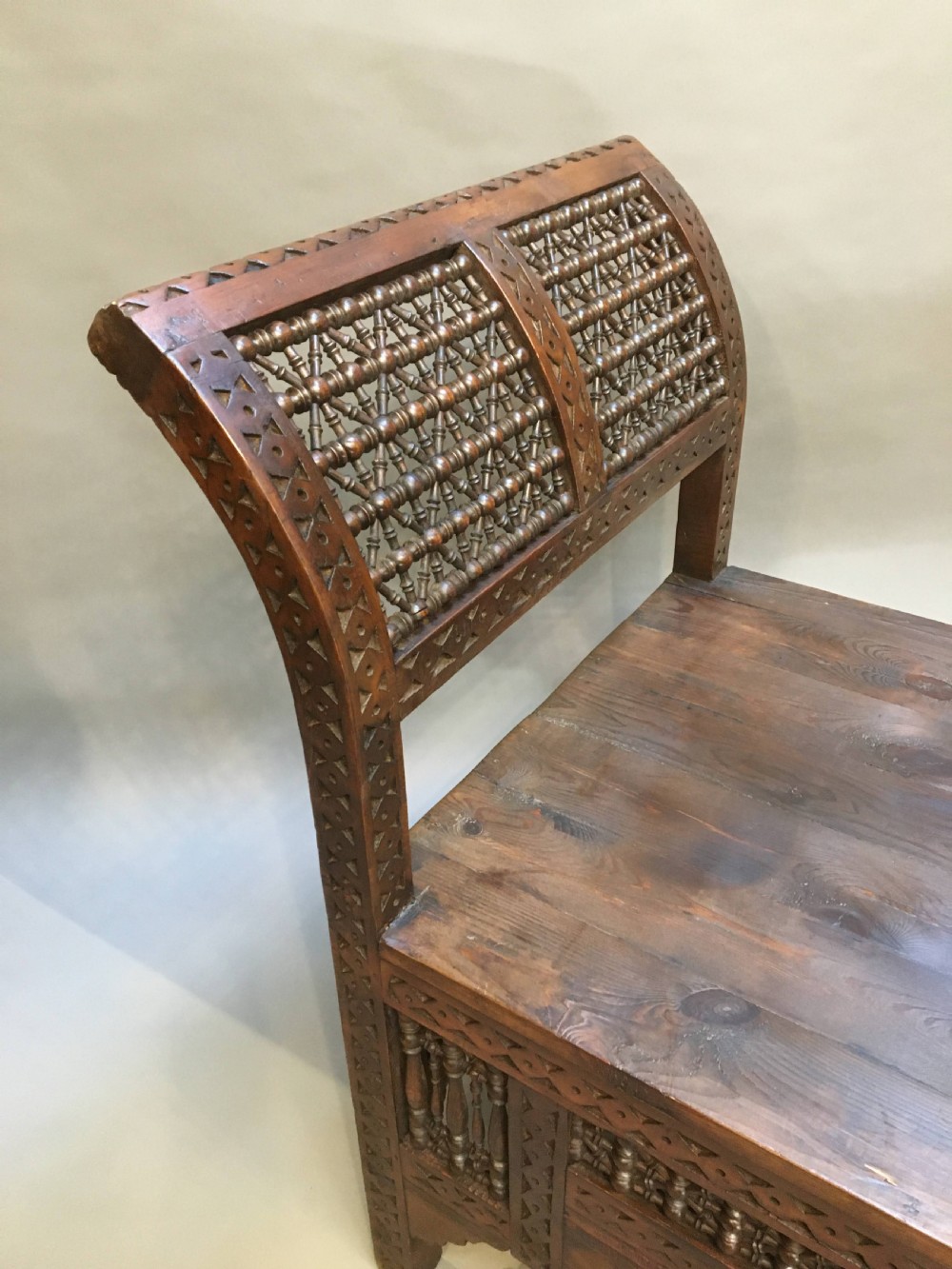 A dark brown carved wooden window seat featuring raised curved arms and geometric mashrabiya lattice panels along the base. View V