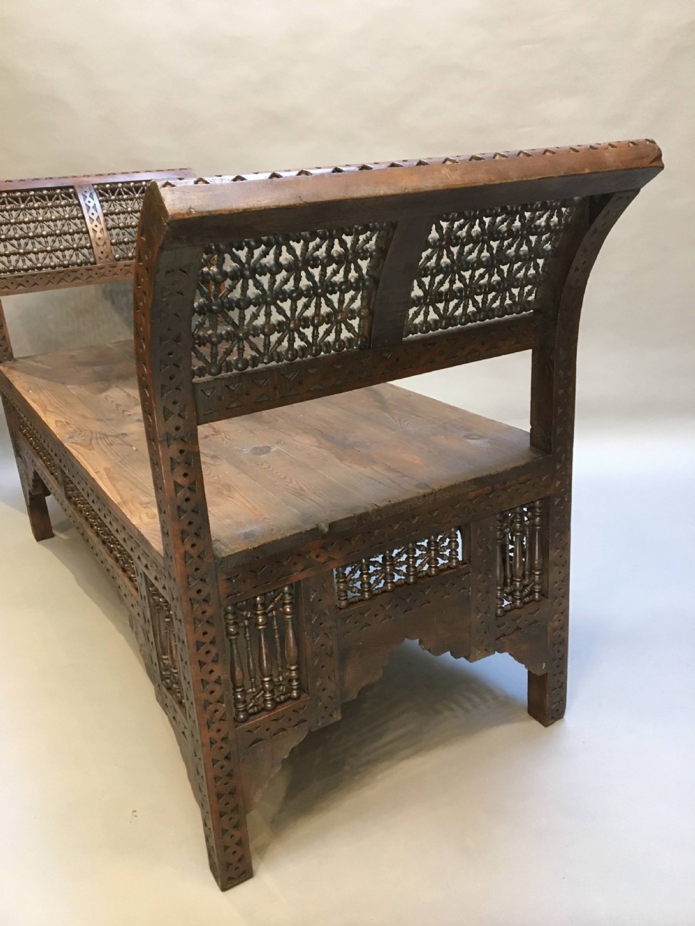 A dark brown carved wooden window seat featuring raised curved arms and geometric mashrabiya lattice panels along the base. View VI