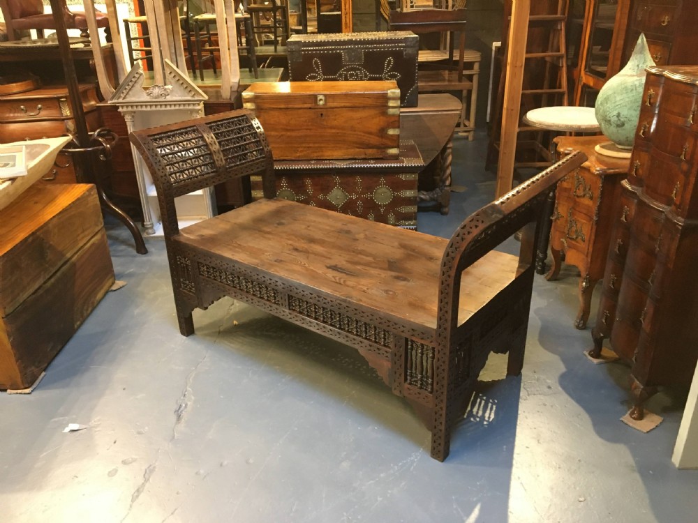 A dark brown carved wooden window seat featuring raised curved arms and geometric mashrabiya lattice panels along the base. View VII