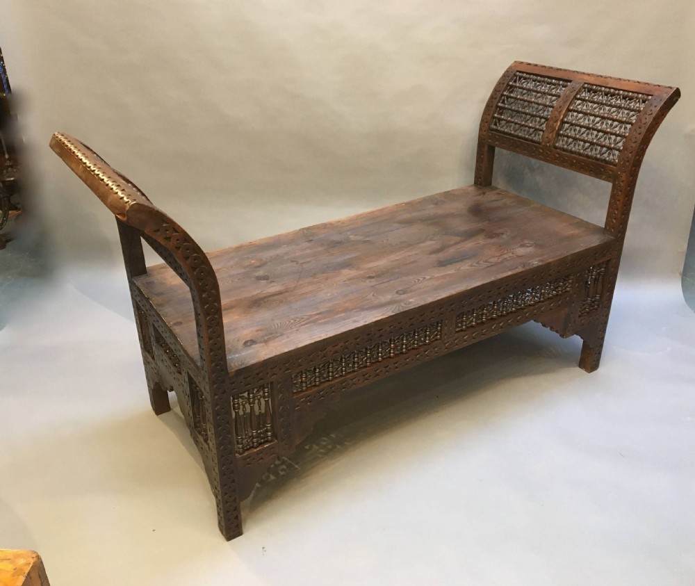 A dark brown carved wooden window seat featuring raised curved arms and geometric mashrabiya lattice panels along the base. View X