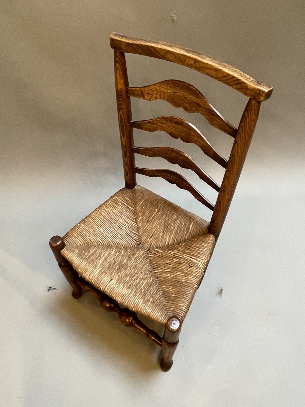 A wooden country side chair featuring a ladderback with four wavy horizontal slats, turned front legs, and a woven rush seat. View VI