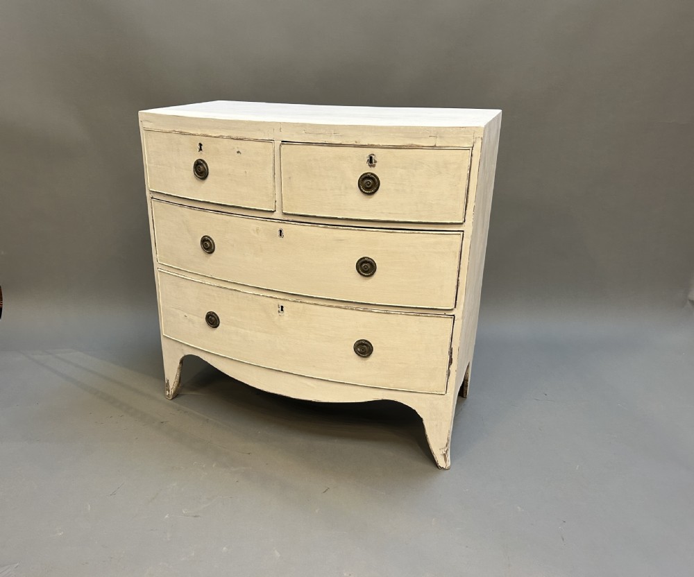 An antique off-white painted bow-front chest of drawers featuring two short drawers over two long drawers with brass circular ring handles.