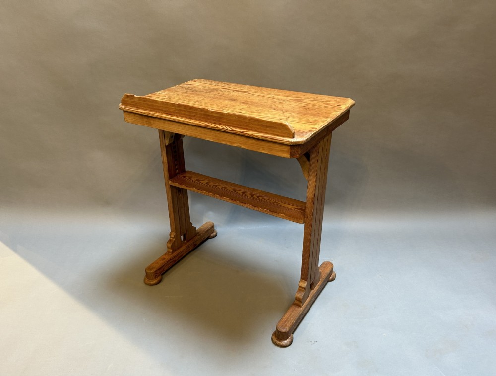 An early twentieth-century pine lectern table featuring a slanted reading surface with a retaining lip, supported by a trestle base with a lower shelf.