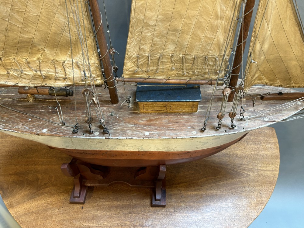A large vintage wooden pond yacht with a two-masted schooner rig, aged sails, and a white and brown painted hull mounted on a dark wooden stand. View III