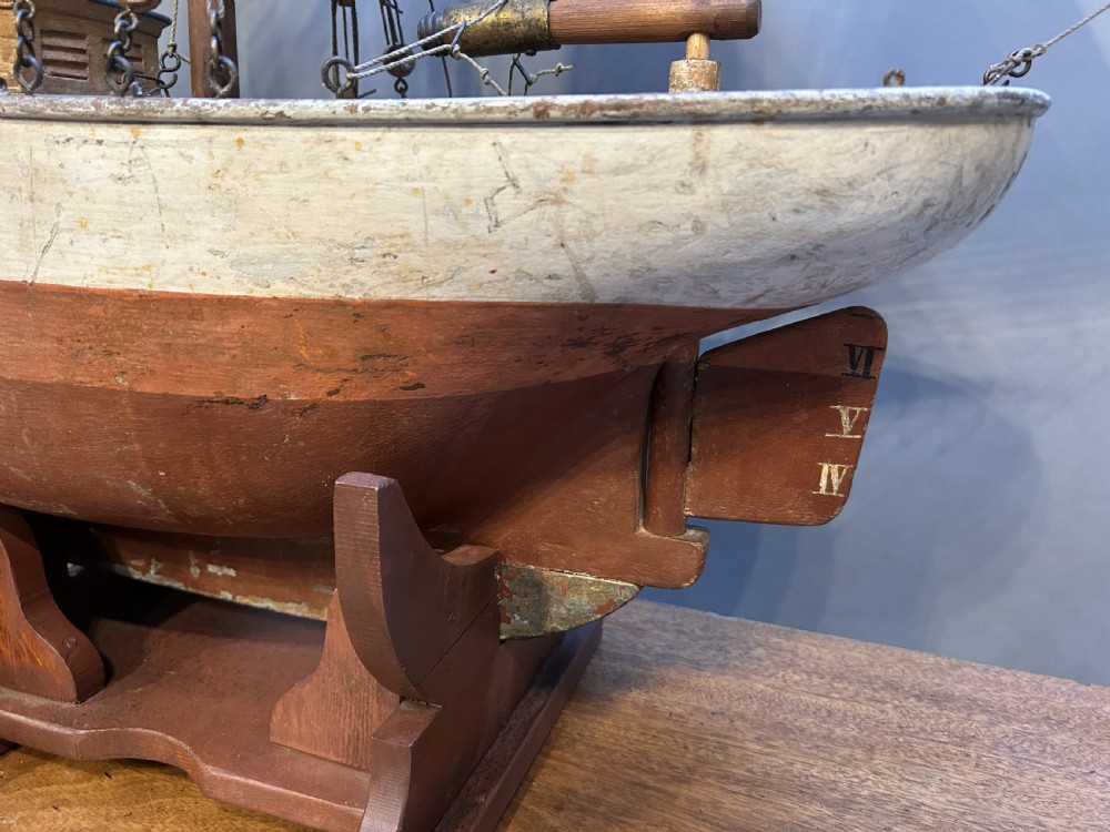 A large vintage wooden pond yacht with a two-masted schooner rig, aged sails, and a white and brown painted hull mounted on a dark wooden stand. View V