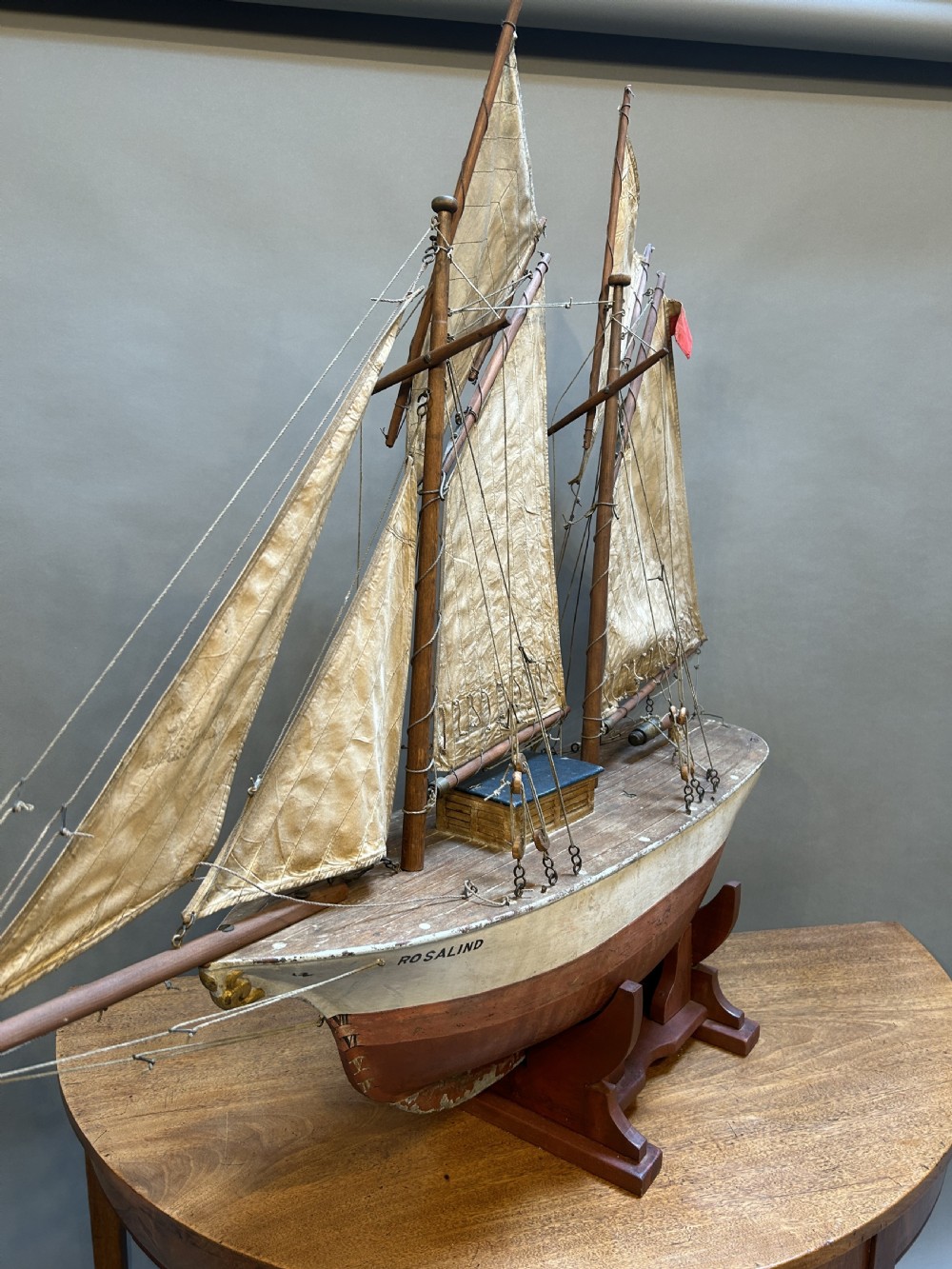 A large vintage wooden pond yacht with a two-masted schooner rig, aged sails, and a white and brown painted hull mounted on a dark wooden stand. View VIII