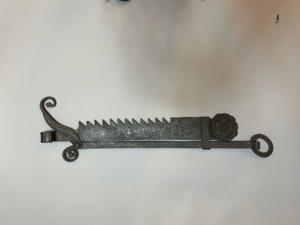 An 18th-century hand-forged wrought iron adjustable chimney crane featuring a sawtooth trammel mechanism, scrolled ends, and a decorative rosette.