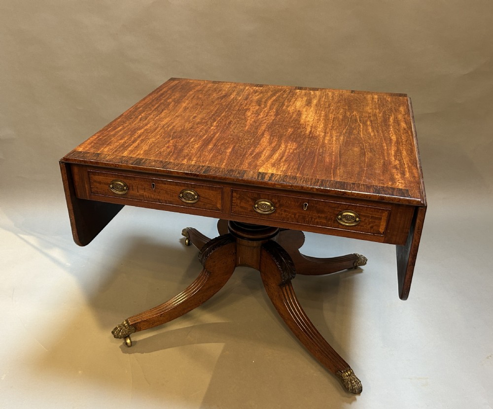A George IV flame mahogany sofa table featuring drop-leaf sides, frieze drawers with brass handles, and a quadruped pedestal base with reeded legs on brass lion paw castors.