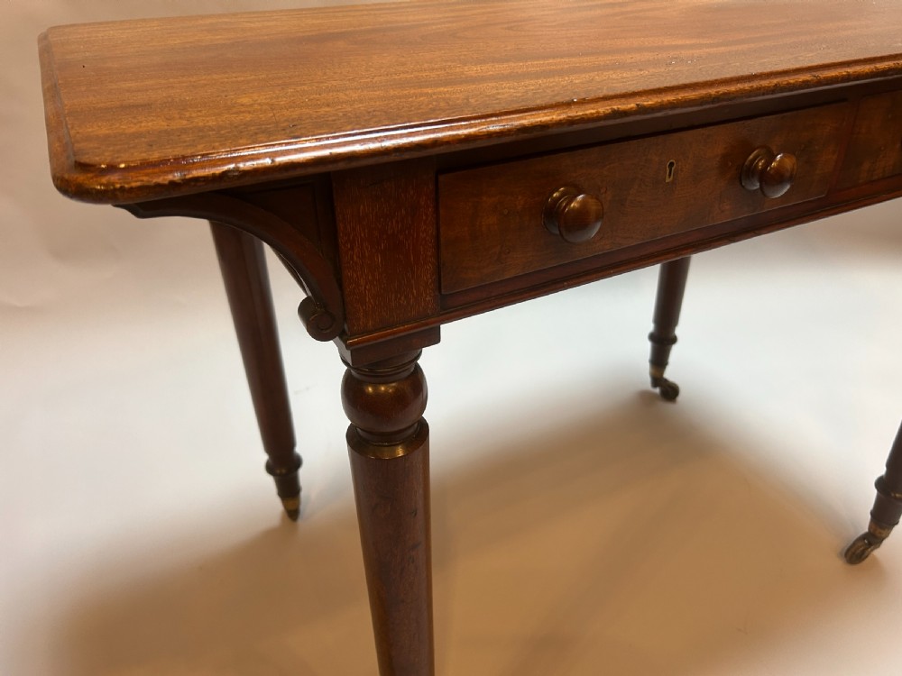 A Victorian mahogany writing table featuring two frieze drawers with turned wooden knob handles and turned legs supported by brass castors. View VI