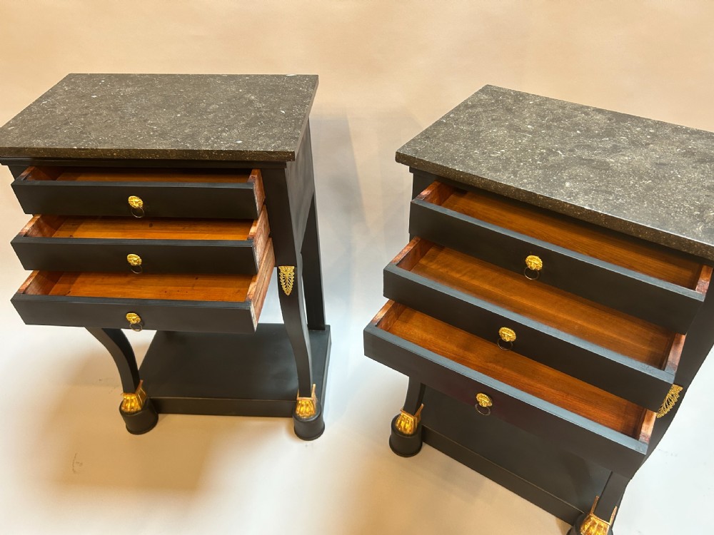 A pair of ebonised black French Empire style bedside chests featuring grey marble tops, three small drawers with ring pulls, curved front legs with gilt paw feet, and an undertier shelf. View V