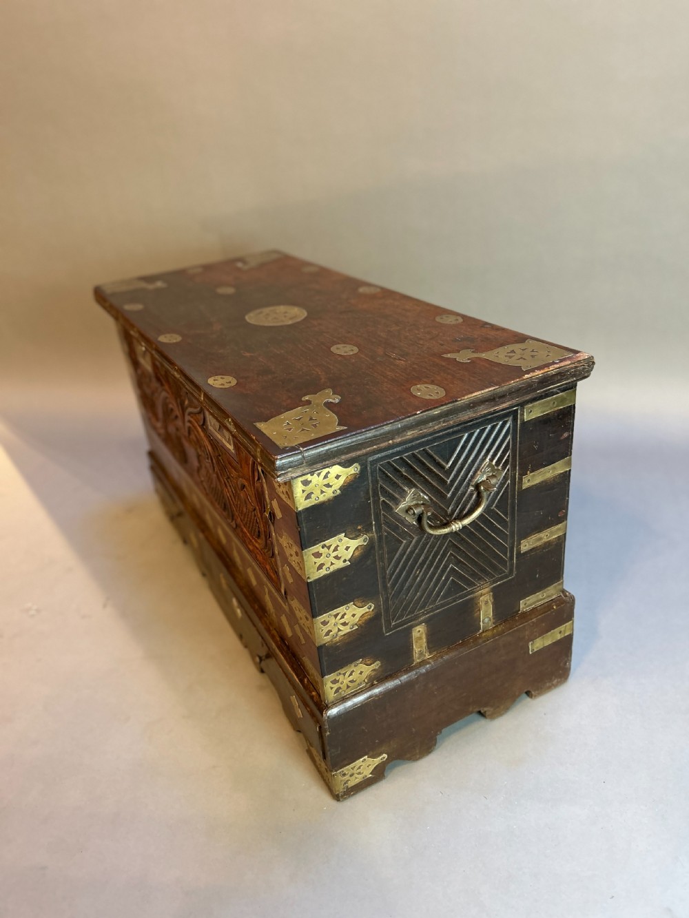 A 19th-century dark hardwood Zanzibar chest featuring intricate pierced brass mountings, studded detailing, a carved foliate front panel, and lower drawers. View V