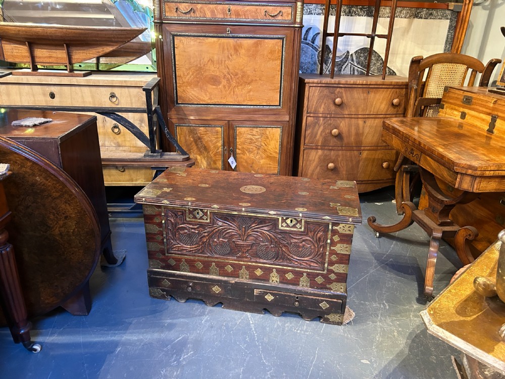 A 19th-century dark hardwood Zanzibar chest featuring intricate pierced brass mountings, studded detailing, a carved foliate front panel, and lower drawers. View VIII