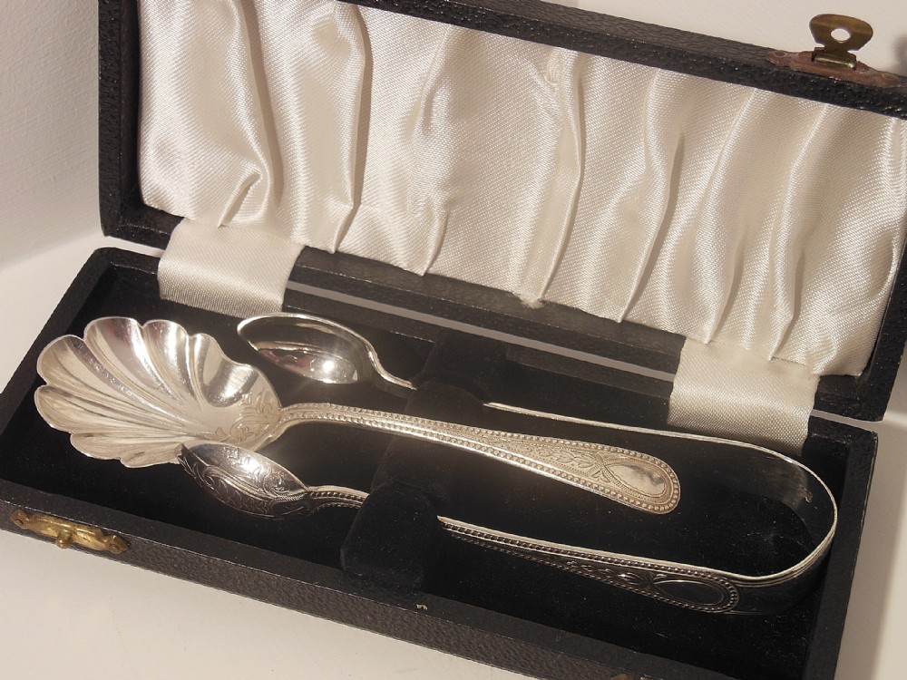 A silver sugar spoon with a shell-shaped bowl alongside matching silver sugar tongs, presented inside a black silk- and velvet-lined box.