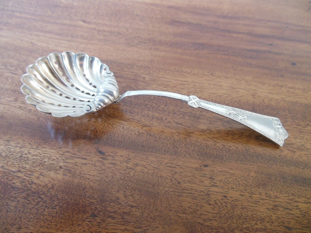 An antique silver sifter spoon featuring a fluted scallop shell bowl with pierced slots and an intricately engraved handle resting on a wooden surface.