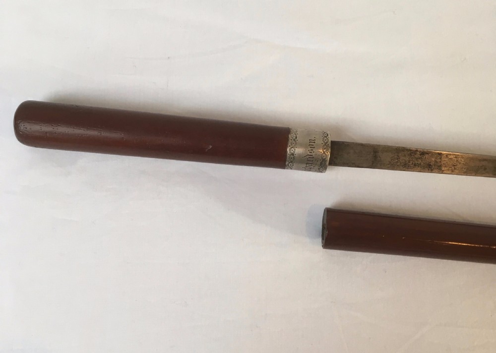 A Victorian hardwood sword stick separated to display a long steel blade beside its dark wooden sheath with a silver collar. View II