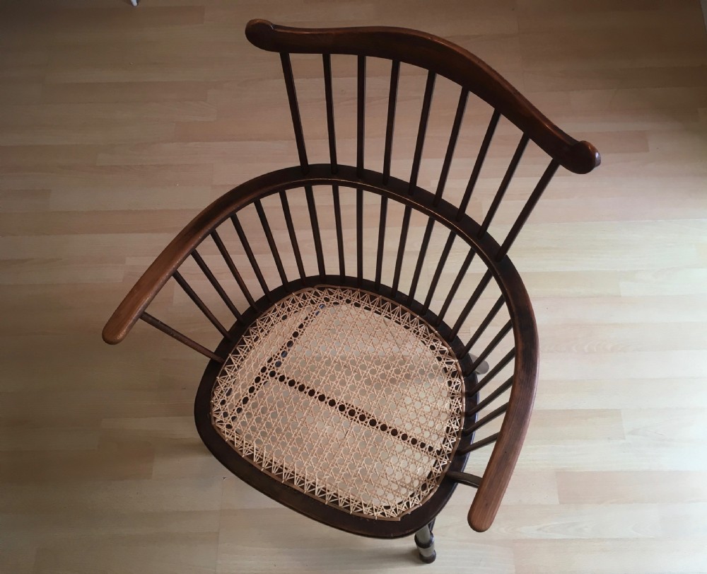 A dark stained wooden stick-back armchair featuring a comb crest rail, spindle uprights, curved armrests, and a woven natural cane seat. View IV