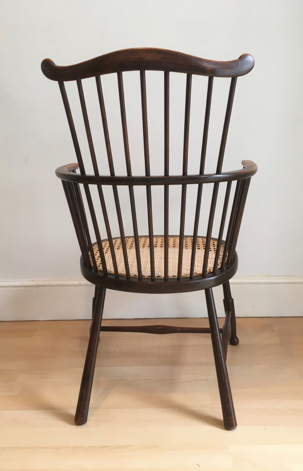 A dark stained wooden stick-back armchair featuring a comb crest rail, spindle uprights, curved armrests, and a woven natural cane seat. View V