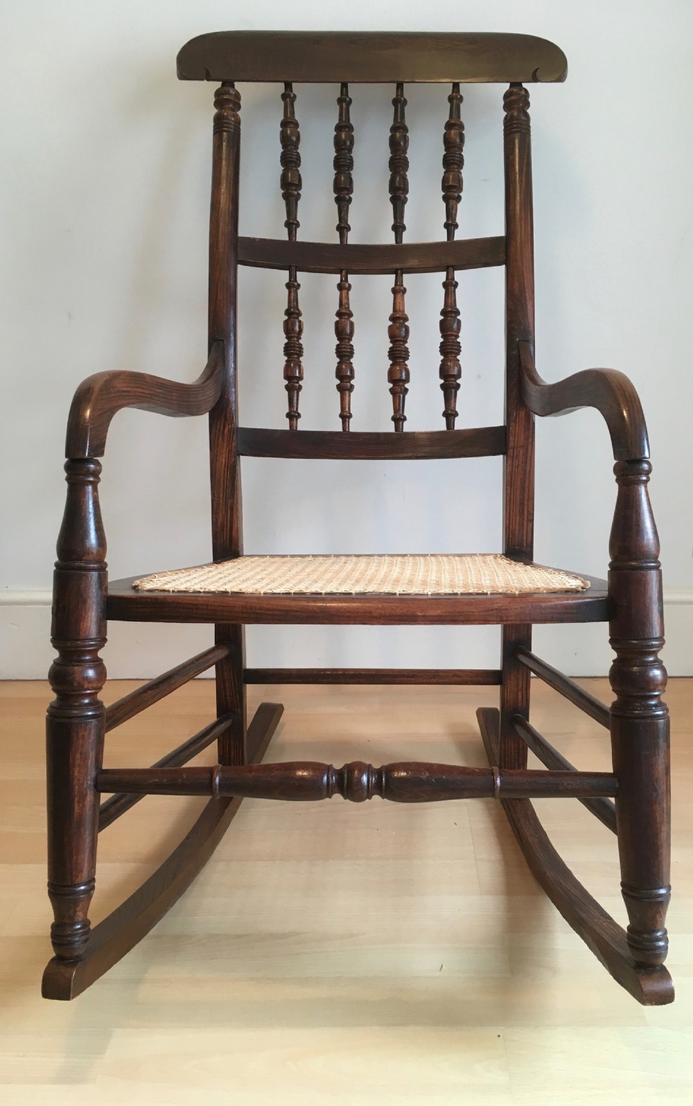 A dark wood 19th-century spindle back rocking chair featuring a woven cane seat, turned spindles, and curved armrests resting on curved runners. View II