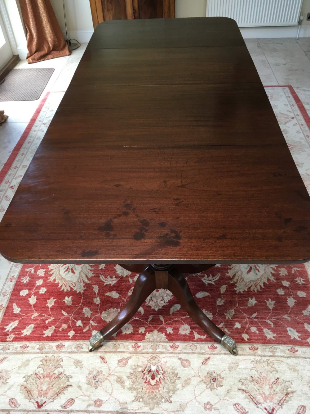 A 19th-century mahogany extendable dining table featuring a polished rectangular top with rounded corners, supported by two turned pillar pedestals on splayed legs with brass castors. View V