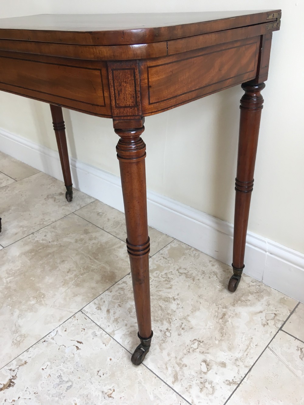 A Regency mahogany fold-over card table featuring turned tapered legs, brass castors, and boxwood stringing inlay along the frieze. View V