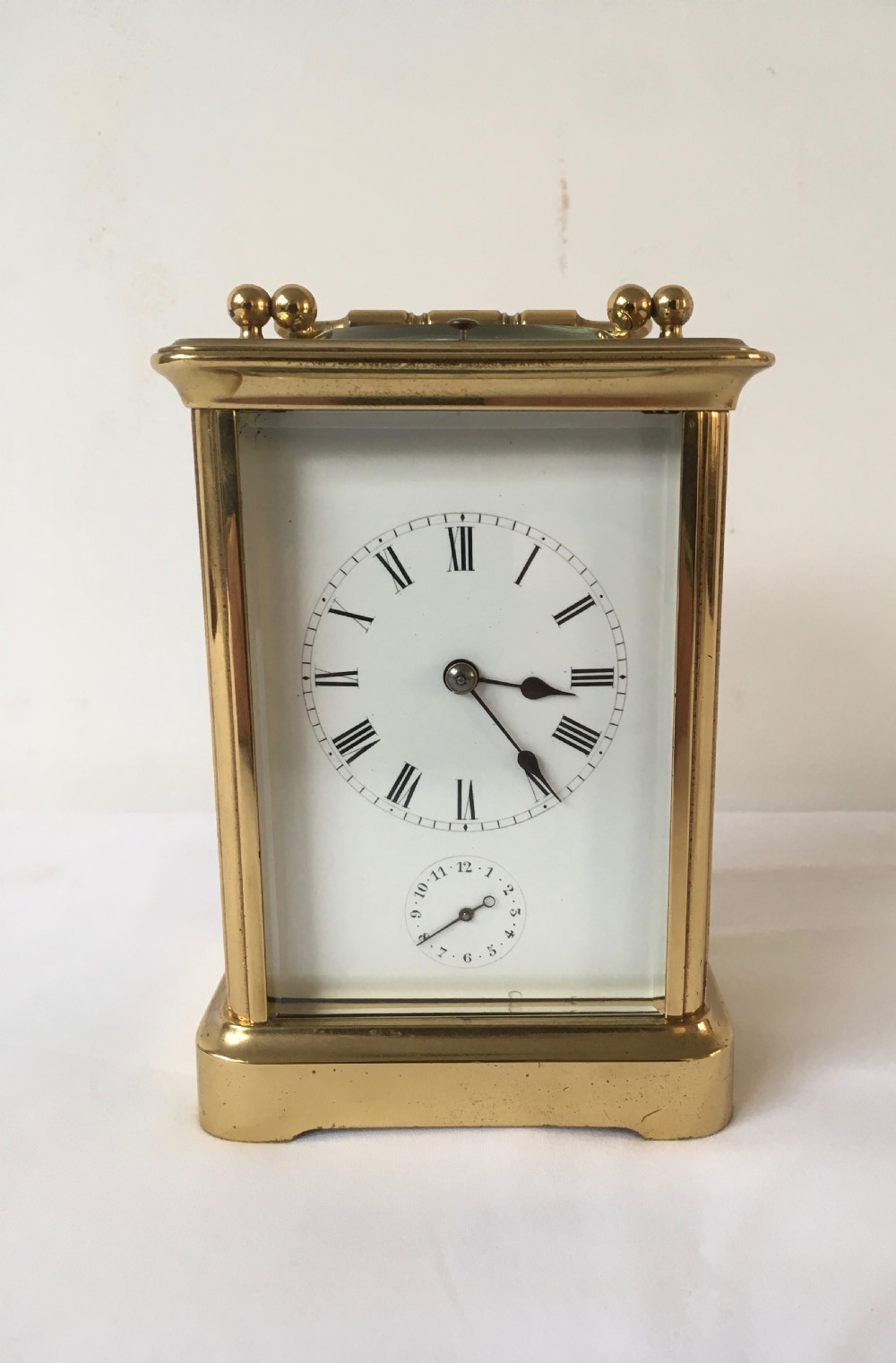 A rectangular brass carriage clock featuring clear bevelled glass panels, a white enamel dial with Roman numerals, a subsidiary alarm dial, and decorative corner finials.