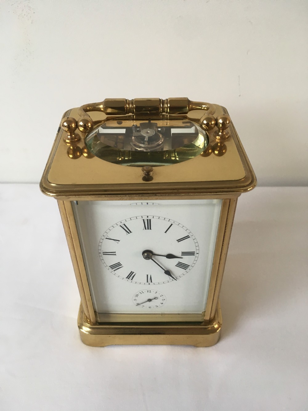 A rectangular brass carriage clock featuring clear bevelled glass panels, a white enamel dial with Roman numerals, a subsidiary alarm dial, and decorative corner finials. View II
