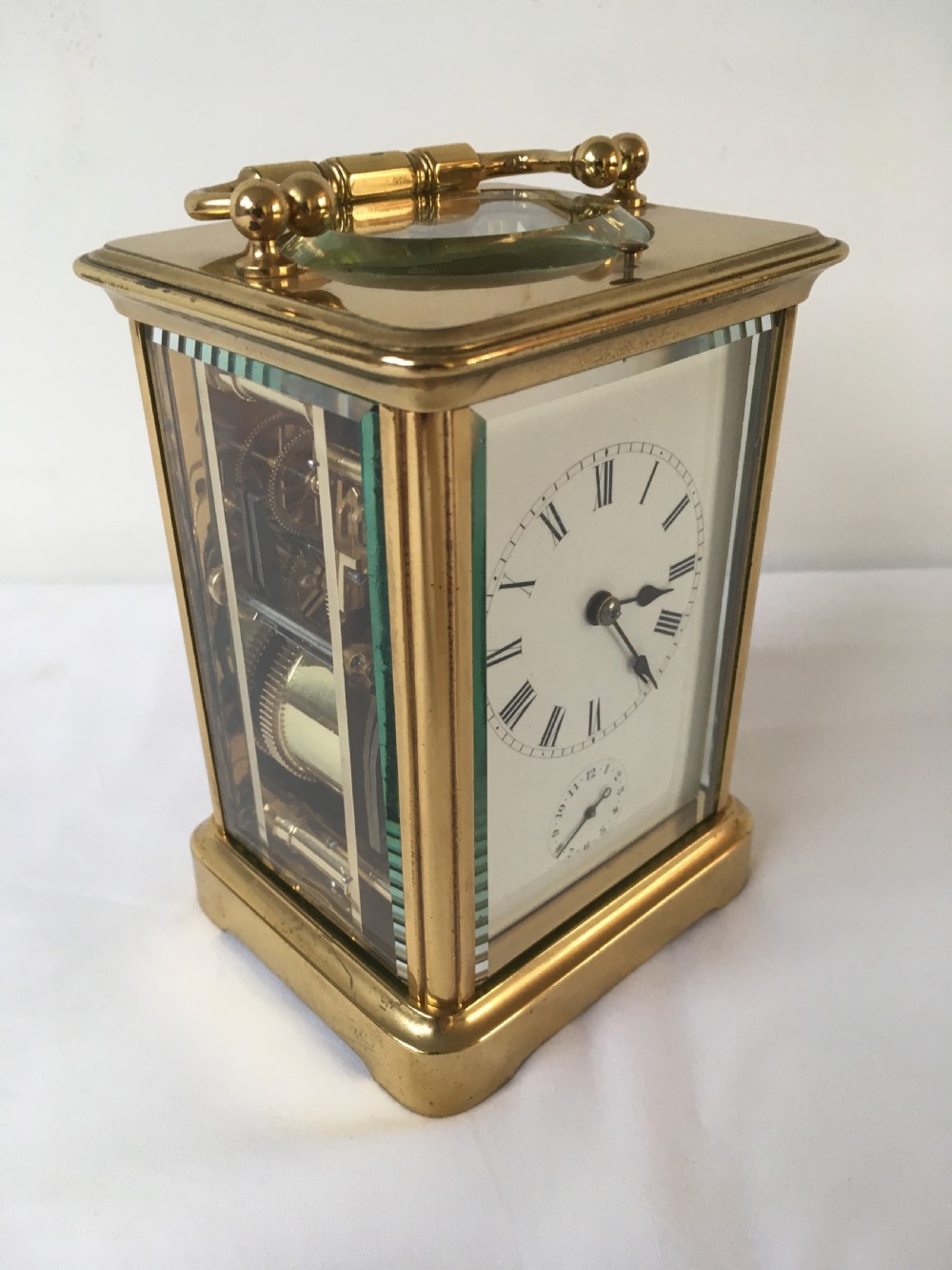 A rectangular brass carriage clock featuring clear bevelled glass panels, a white enamel dial with Roman numerals, a subsidiary alarm dial, and decorative corner finials. View III