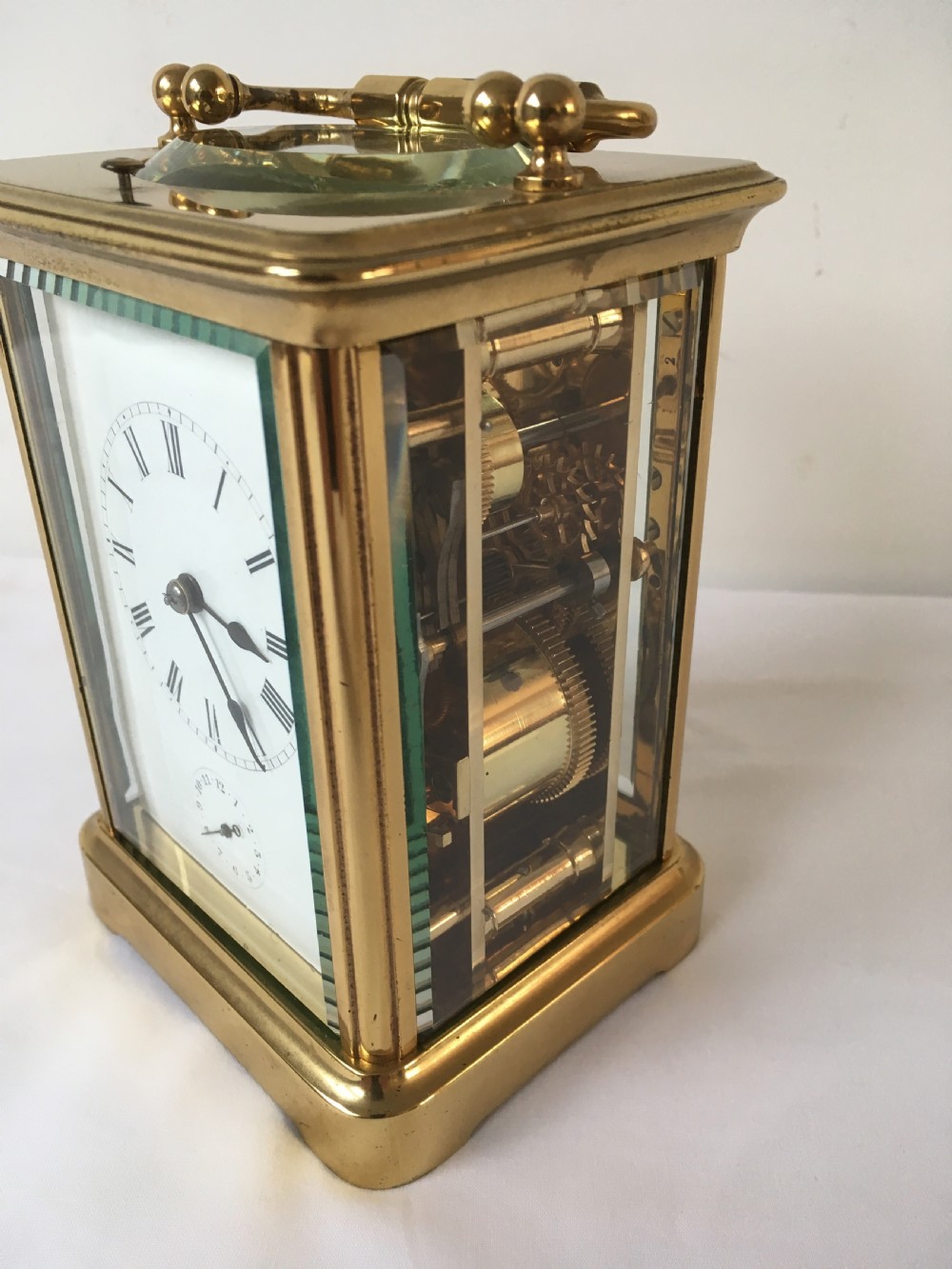 A rectangular brass carriage clock featuring clear bevelled glass panels, a white enamel dial with Roman numerals, a subsidiary alarm dial, and decorative corner finials. View IV