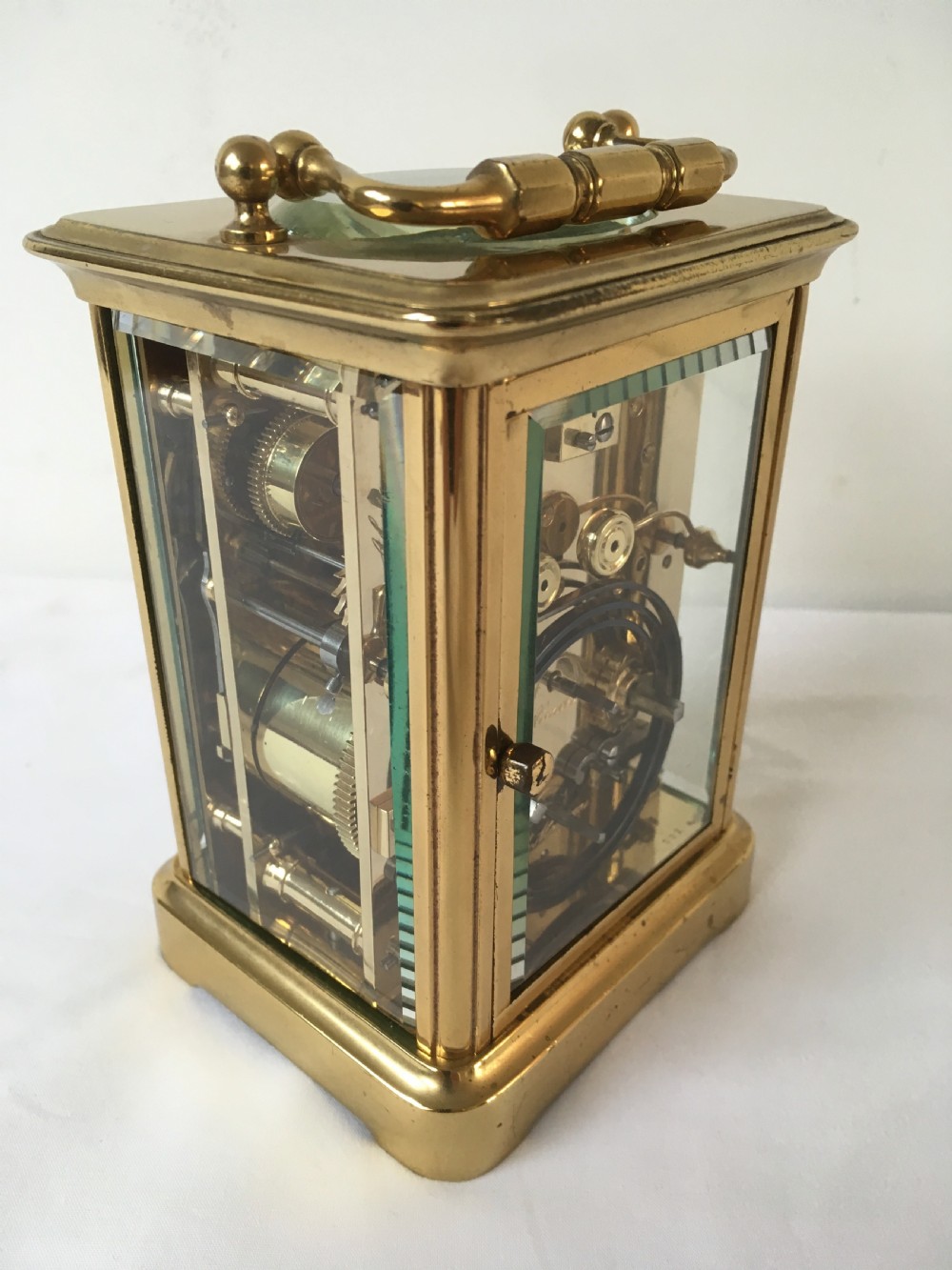 A rectangular brass carriage clock featuring clear bevelled glass panels, a white enamel dial with Roman numerals, a subsidiary alarm dial, and decorative corner finials. View V