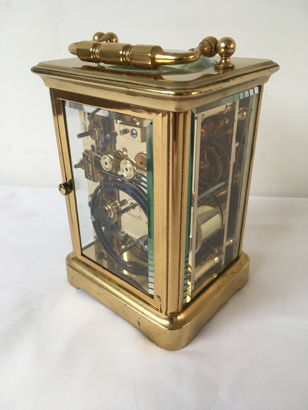 A rectangular brass carriage clock featuring clear bevelled glass panels, a white enamel dial with Roman numerals, a subsidiary alarm dial, and decorative corner finials. View VI