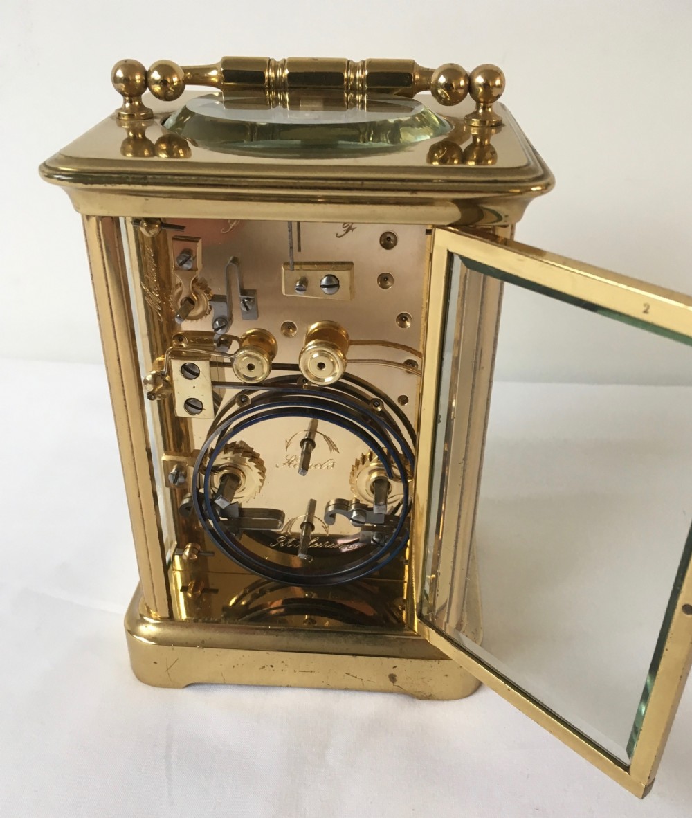 A rectangular brass carriage clock featuring clear bevelled glass panels, a white enamel dial with Roman numerals, a subsidiary alarm dial, and decorative corner finials. View VII
