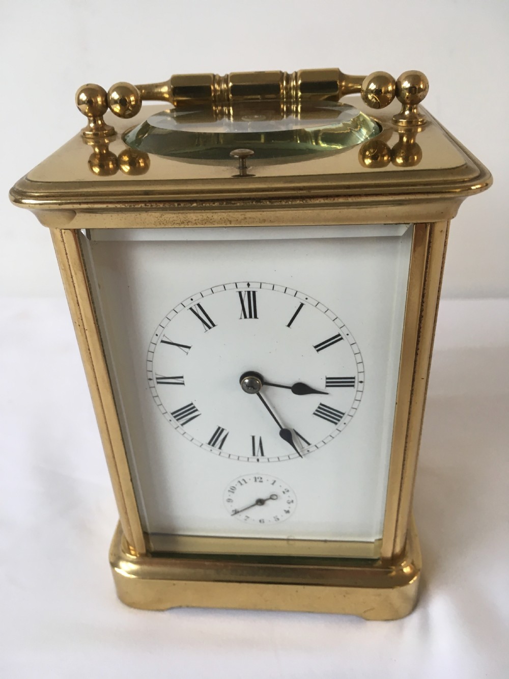 A rectangular brass carriage clock featuring clear bevelled glass panels, a white enamel dial with Roman numerals, a subsidiary alarm dial, and decorative corner finials. View IX