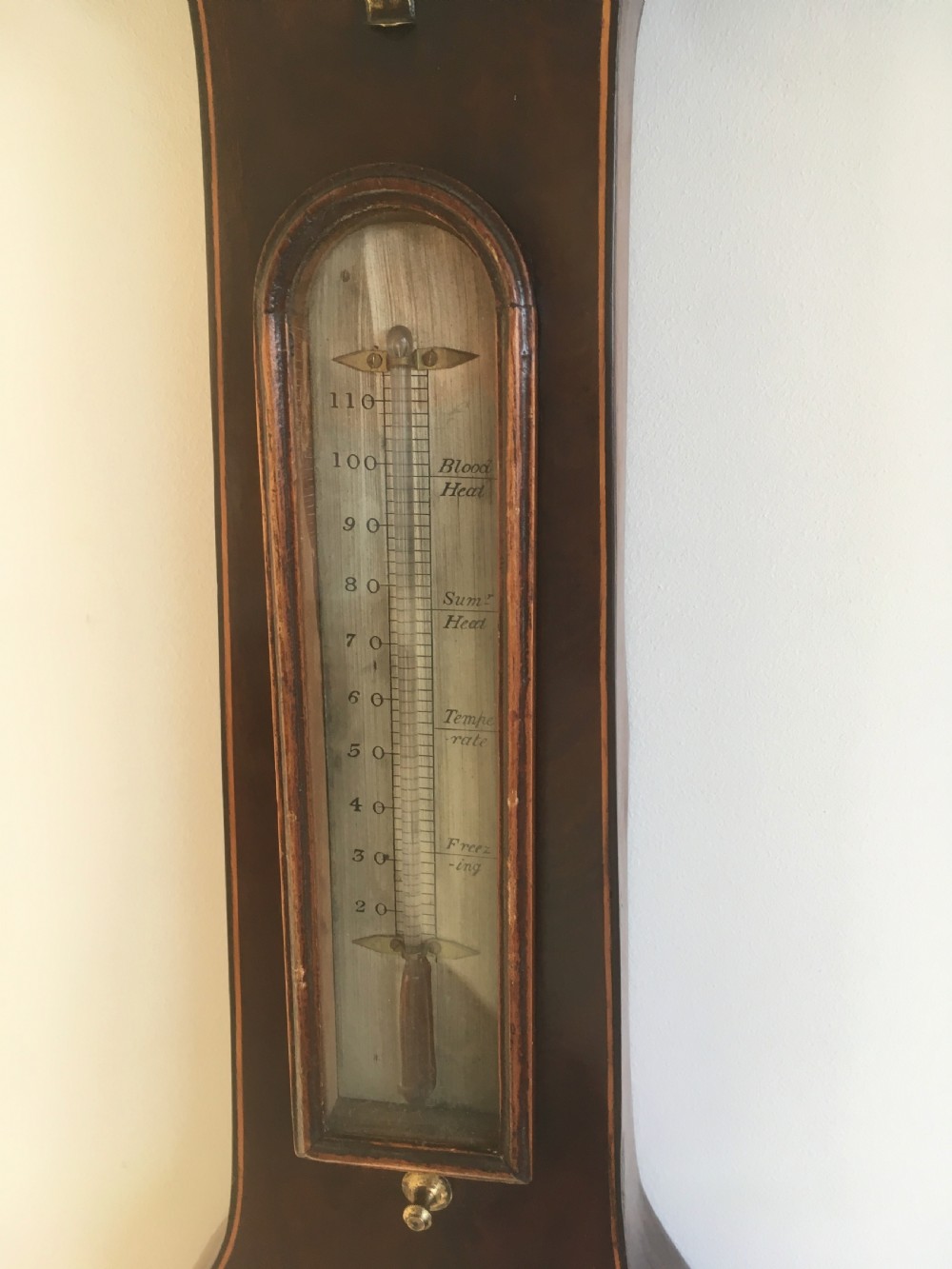 An antique mahogany banjo wheel barometer featuring a top brass finial, thermometer display, central convex mirror, and silvered register dial. View III