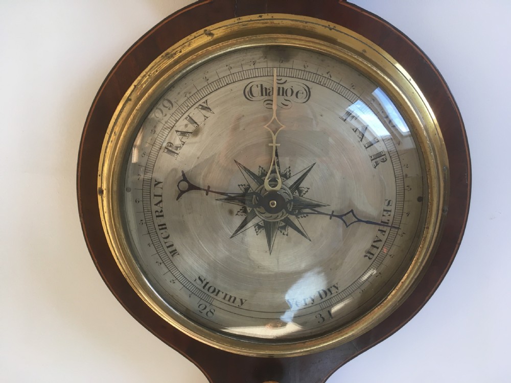 An antique mahogany banjo wheel barometer featuring a top brass finial, thermometer display, central convex mirror, and silvered register dial. View V