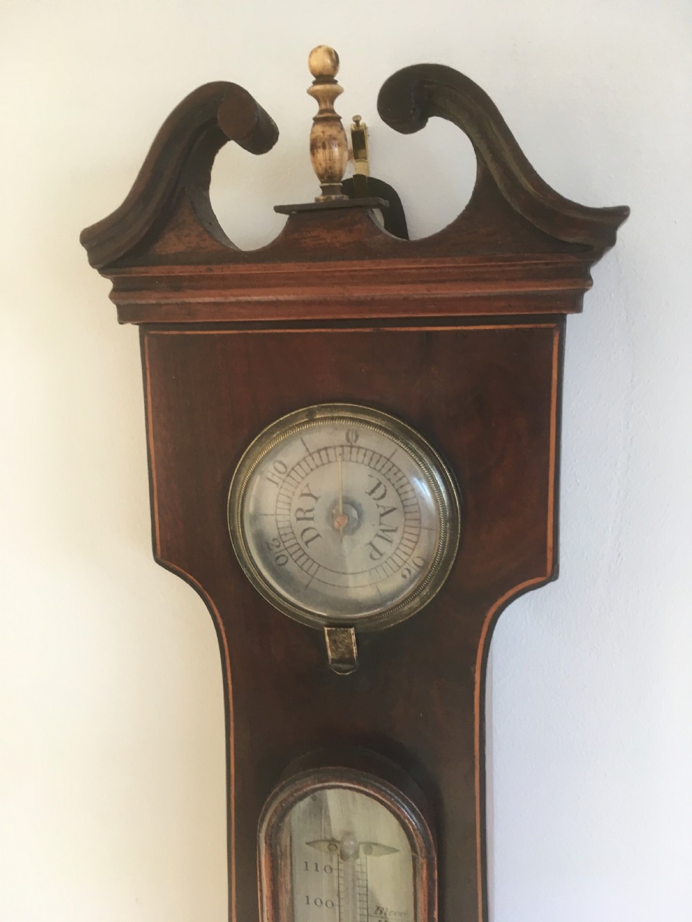 An antique mahogany banjo wheel barometer featuring a top brass finial, thermometer display, central convex mirror, and silvered register dial. View VII