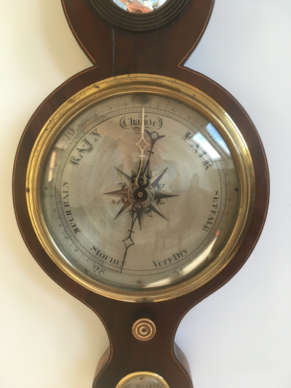 An antique mahogany banjo wheel barometer featuring a top brass finial, thermometer display, central convex mirror, and silvered register dial. View IX