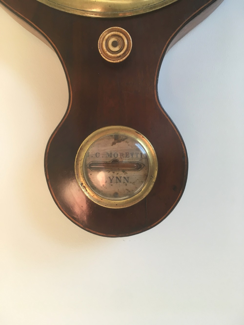 An antique mahogany banjo wheel barometer featuring a top brass finial, thermometer display, central convex mirror, and silvered register dial. View X