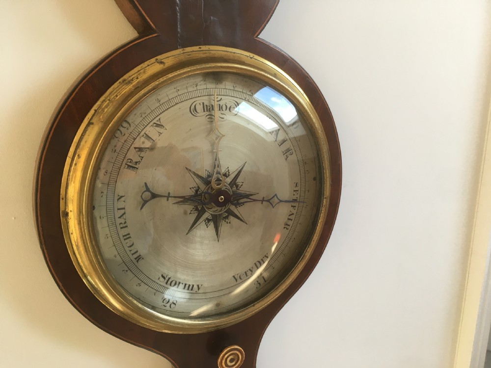 An antique mahogany banjo wheel barometer featuring a top brass finial, thermometer display, central convex mirror, and silvered register dial. View XII