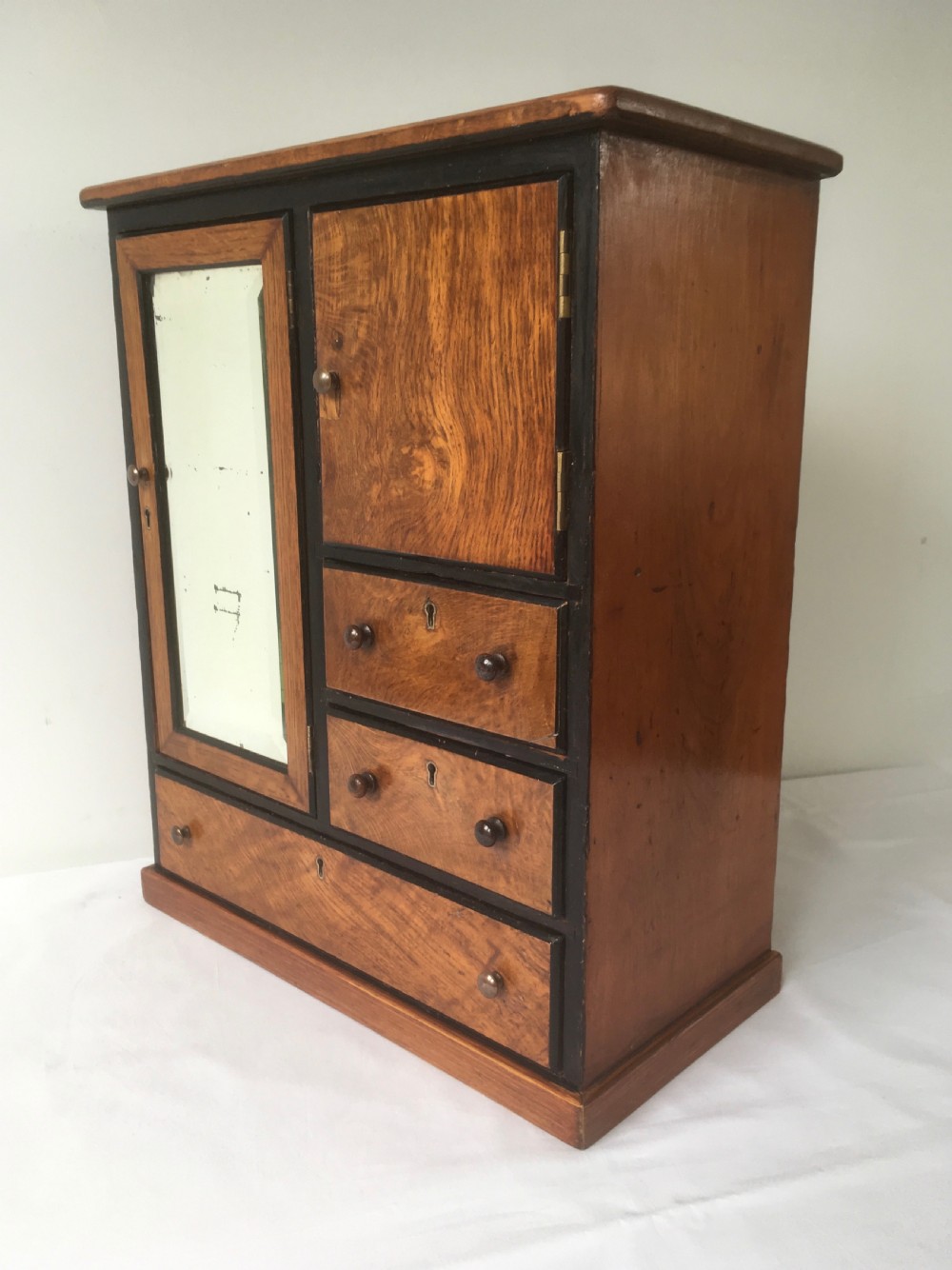 A miniature oak compactum wardrobe featuring a left door with a rectangular mirror, a right cupboard door, three small drawers, wooden knob handles, and dark ebonised framing.