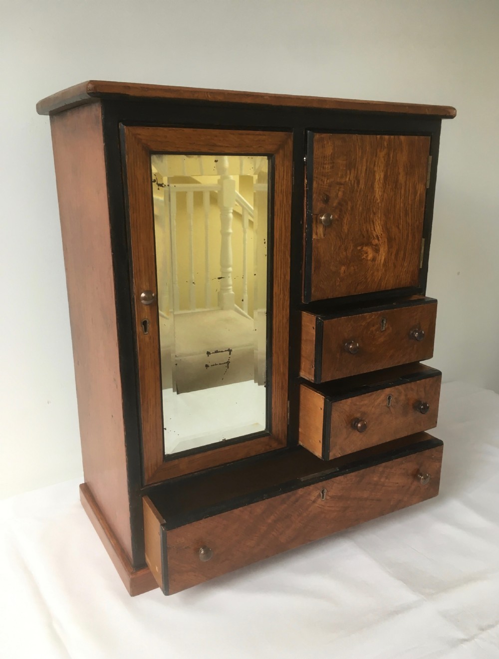 A miniature oak compactum wardrobe featuring a left door with a rectangular mirror, a right cupboard door, three small drawers, wooden knob handles, and dark ebonised framing. View II