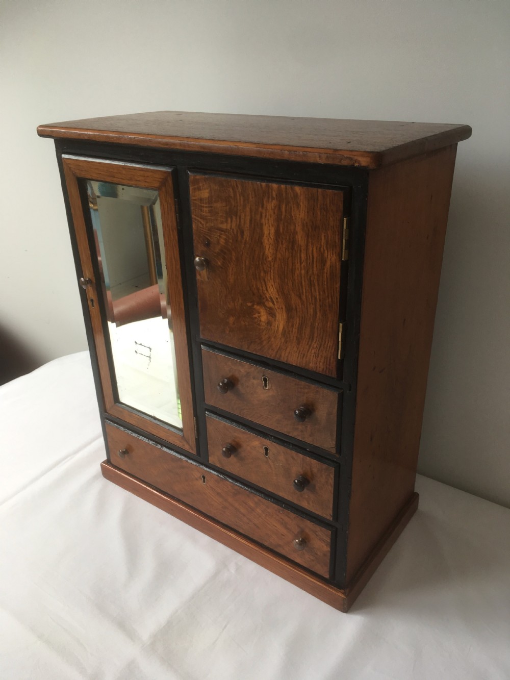 A miniature oak compactum wardrobe featuring a left door with a rectangular mirror, a right cupboard door, three small drawers, wooden knob handles, and dark ebonised framing. View III