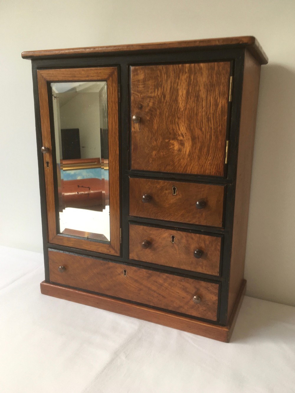 A miniature oak compactum wardrobe featuring a left door with a rectangular mirror, a right cupboard door, three small drawers, wooden knob handles, and dark ebonised framing. View VII