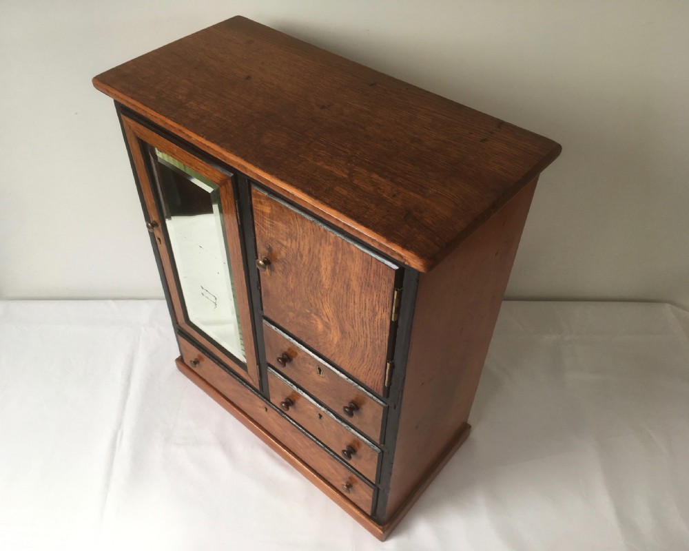 A miniature oak compactum wardrobe featuring a left door with a rectangular mirror, a right cupboard door, three small drawers, wooden knob handles, and dark ebonised framing. View XII