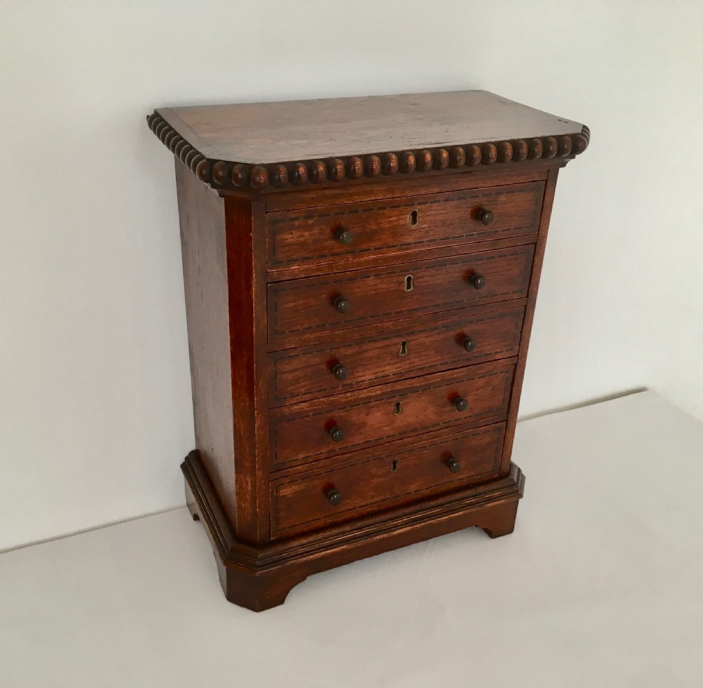 A small dark oak tabletop collectors cabinet featuring five inlaid drawers, turned wooden knobs, and a carved beaded top edge.