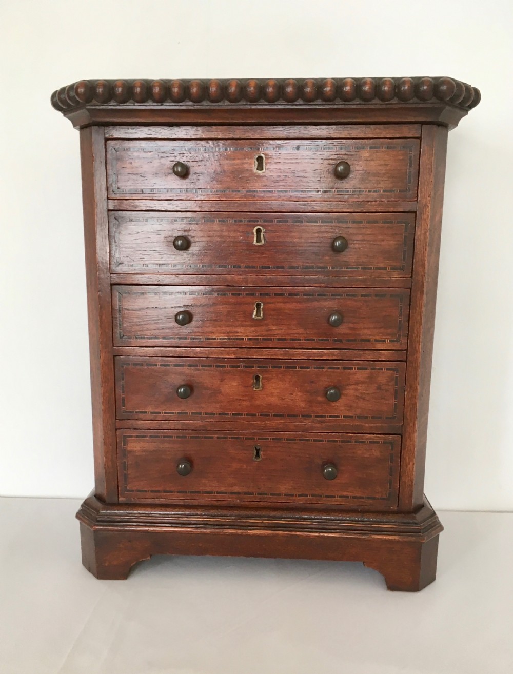 A small dark oak tabletop collectors cabinet featuring five inlaid drawers, turned wooden knobs, and a carved beaded top edge. View V