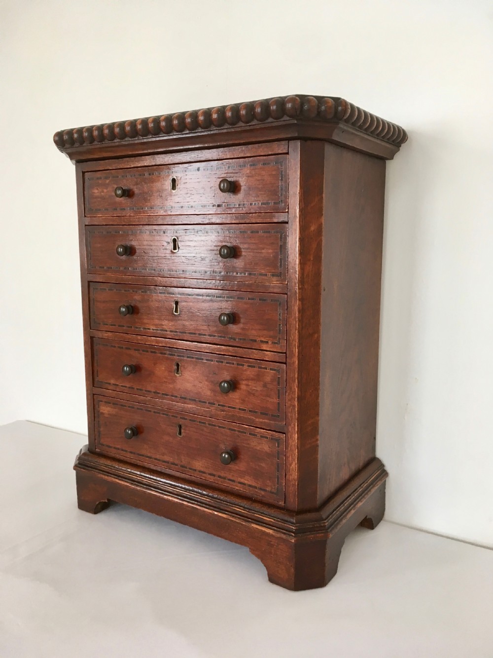 A small dark oak tabletop collectors cabinet featuring five inlaid drawers, turned wooden knobs, and a carved beaded top edge. View VII