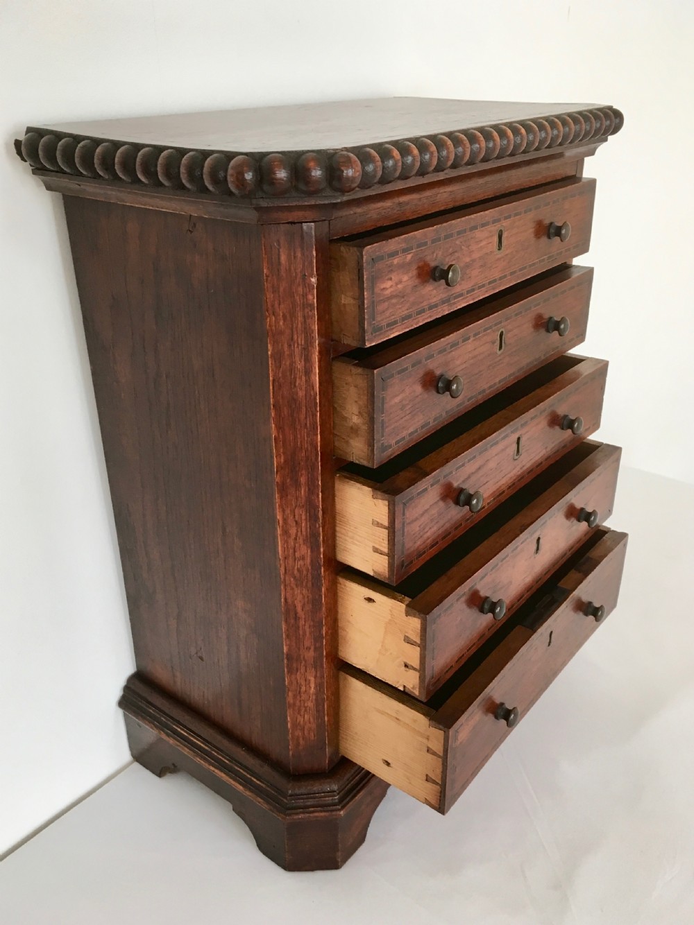 A small dark oak tabletop collectors cabinet featuring five inlaid drawers, turned wooden knobs, and a carved beaded top edge. View XII