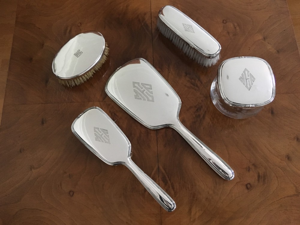 A five-piece silver Art Deco dressing table set featuring a hand mirror, brushes, and a silver-topped glass pot, each detailed with geometric engraved monograms.